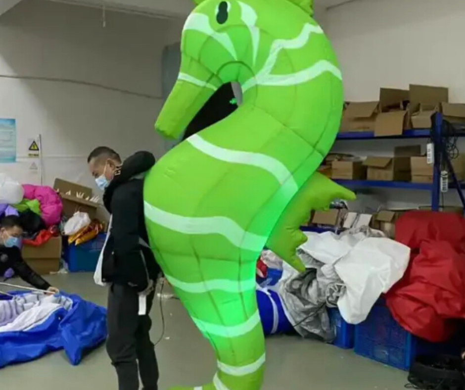 Person wearing a green inflatable seahorse costume in an indoor setting with boxes and bags.
