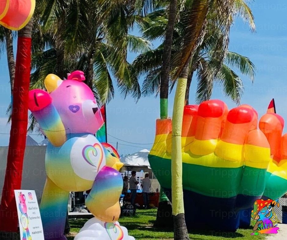 Inflatable rainbow-colored bear and other shapes with palm trees and a clear sky in the background.