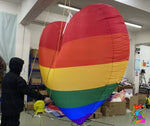 Person working with a large rainbow-colored heart-shaped kite in an indoor setting.