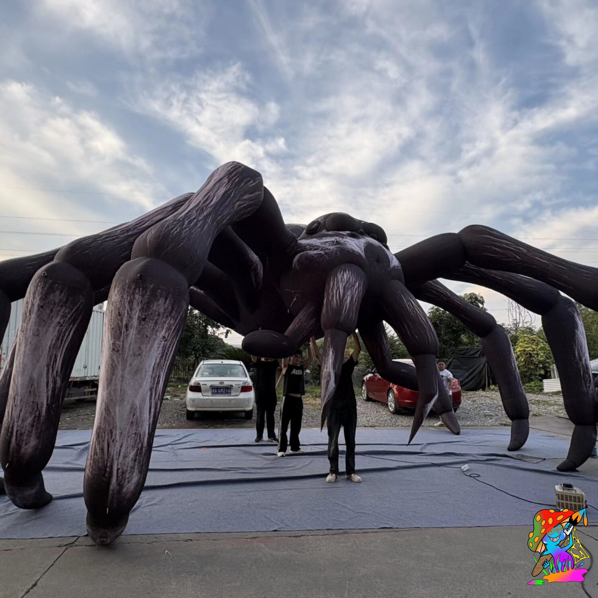A giant inflatable tarantula-like spider sculpture dominates an outdoor paved area, with several people standing underneath it for scale. The sculpture is dark charcoal/black with weathered gray-brown shading.