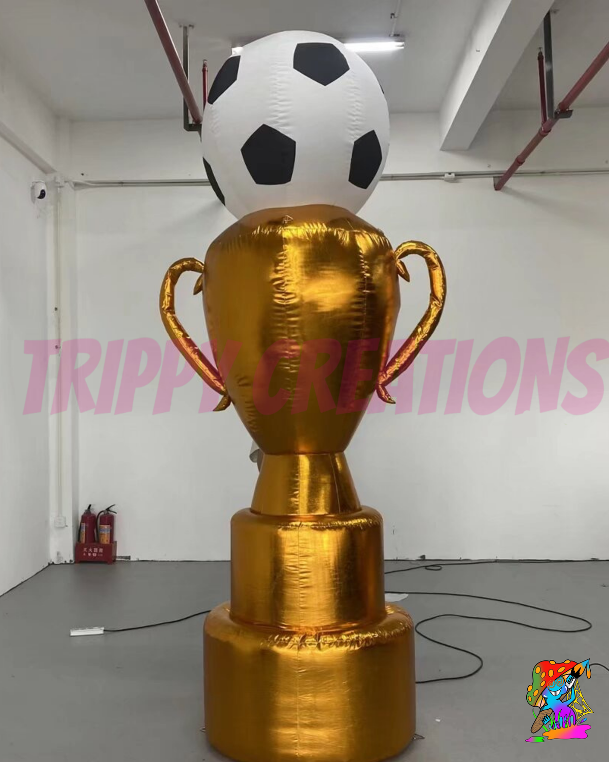 A large inflatable trophy inflatable with a shiny gold finish and a black-and-white soccer ball on top, photographed in a warehouse-like indoor space. A pink 'TRIPY CREATIONS' watermark overlays the center, and a small Trippy Creations logo appears in the bottom-right corner.