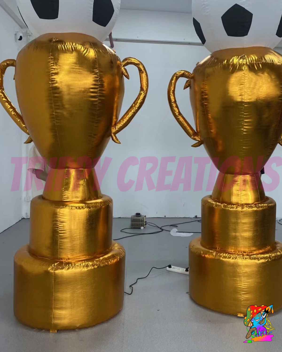 Two large inflatable gold trophy cups displayed indoors on a gray floor, with a soccer ball balloon partially visible behind them. A pink 'TRIPSY CREATIONS' watermark overlays the center.