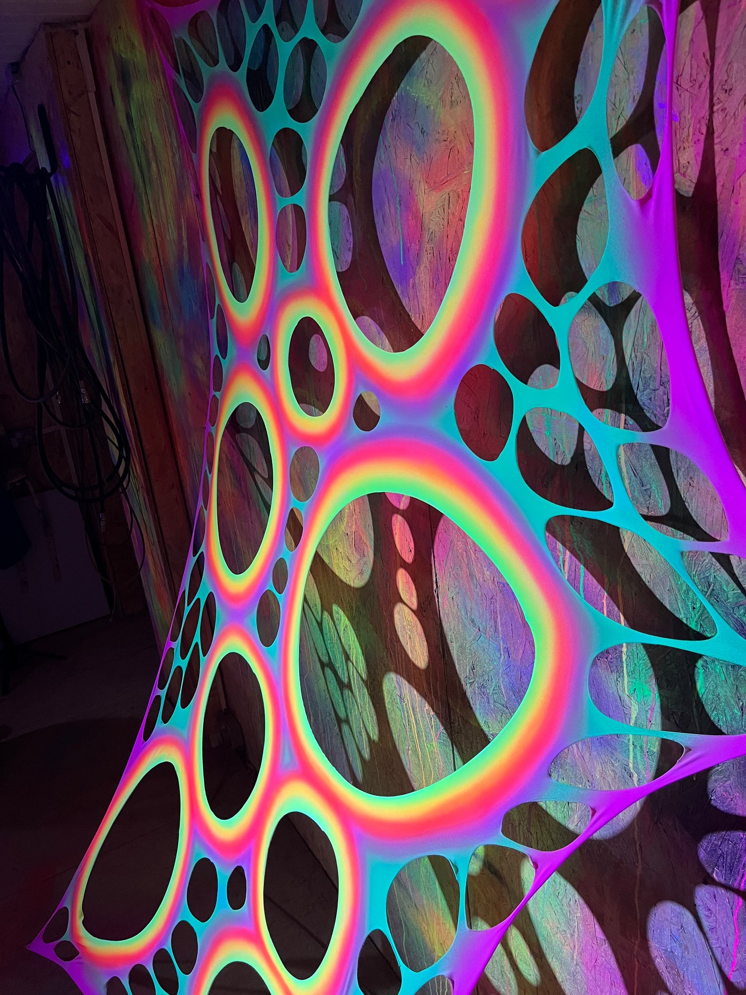 A neon, psychedelic wall hanging or tapestry with a large organic bubble pattern in glowing rings and web-like outlines, photographed indoors against a dark room background.