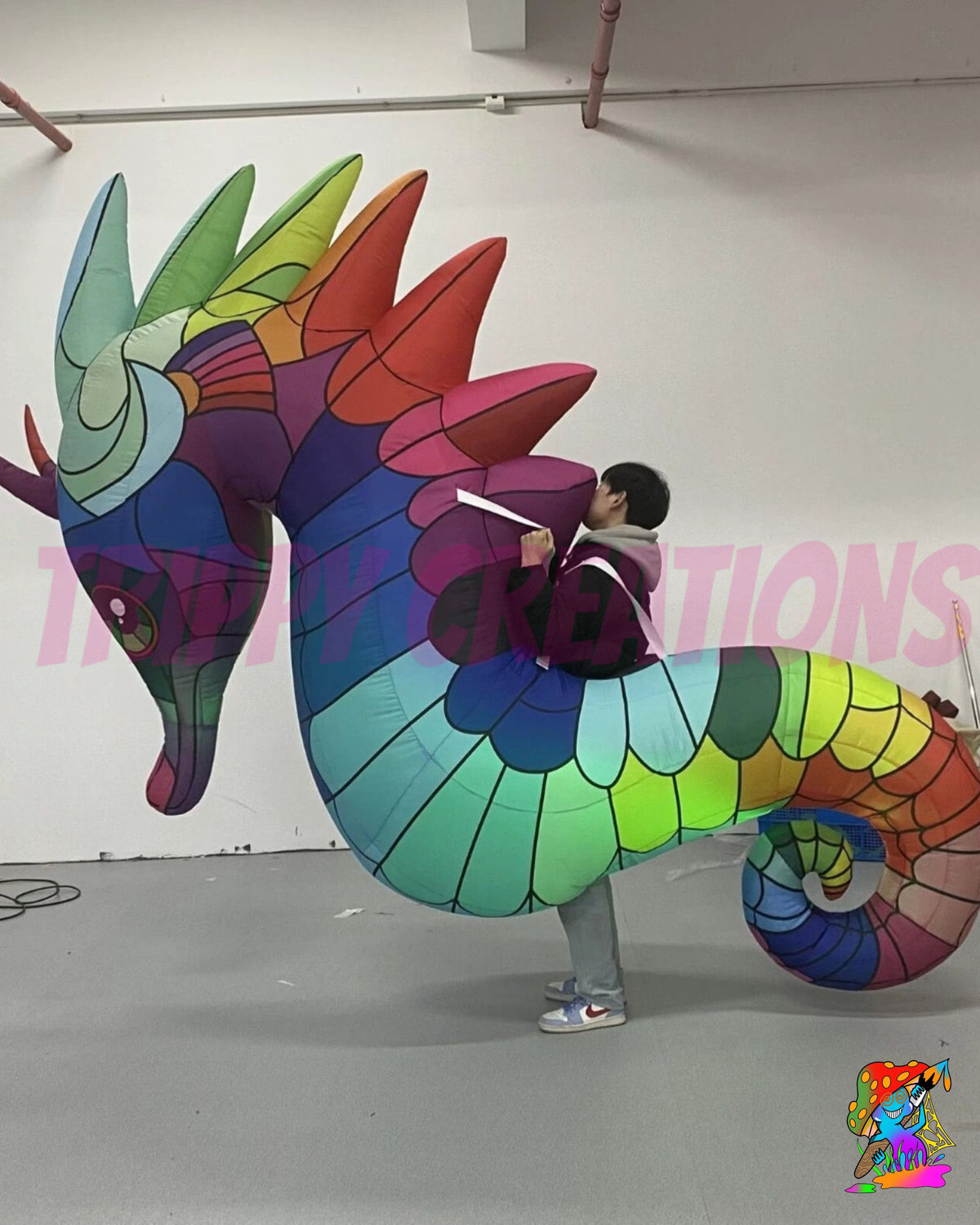 A person wearing a large, colorful inflatable seahorse costume stands indoors against a white wall. The costume features a rainbow of colors including green, blue, purple, red, orange, and yellow in a mosaic pattern.